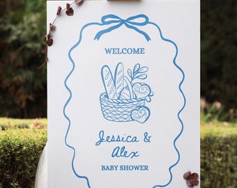Puede incluir: Un letrero blanco sobre un caballete de madera con texto e ilustraciones azules. El letrero dice "WELCOME" encima de una cesta con pan y vegetación, y "Jessica & Alex BABY SHOWER" debajo. Ramas de eucalipto decoran los bordes.
