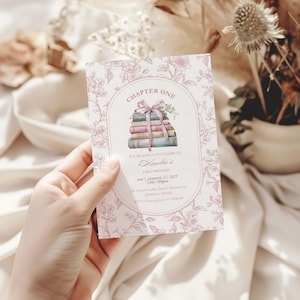 May include: A pink floral invitation card with the text "Chapter One" and "Amelia's First Birthday". The card features a watercolor illustration of a stack of books tied with a ribbon. The card is held by a hand.