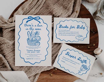 Puede incluir: Invitaciones y tarjetas de baby shower con texto azul sobre papel blanco. La invitación principal dice "there's a bun in the oven" con un gráfico de cesta. Otras tarjetas dicen "Books for Baby" y "Diaper Raffle". Las tarjetas están sobre una tabla de madera con un paño beige.