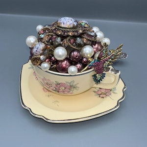 May include: A decorative teacup and saucer set filled with pearls and a large brooch. The teacup is cream-colored with pink floral designs. A jeweled hummingbird is perched on the rim. The brooch is a mix of purple and clear stones.