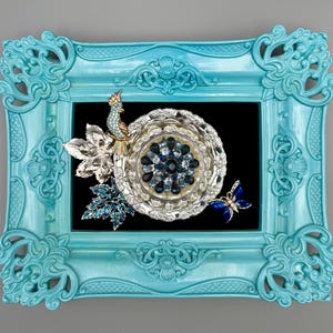 May include: A turquoise ornate picture frame with a black background. The frame holds a decorative arrangement featuring a peacock, a silver flower, and a blue butterfly. The arrangement includes a silver and blue circular design.