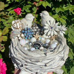 May include: A decorative bird nest sculpture with two small, gray birds perched on the edge. The nest is filled with pearls, beads, and vintage brooches in various colors. Pink roses and green foliage provide a natural backdrop.