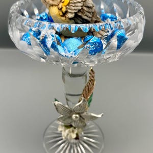 May include: A clear cut-glass pedestal bowl filled with blue foil-wrapped candies. A small, painted bird figurine sits inside the bowl. The glass stem is decorated with a silver-toned metal flower, a small pearl, and a textured leaf accent.