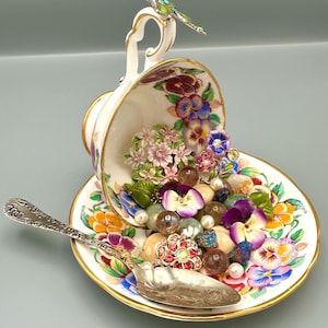 Puede incluir: Un juego decorativo de taza y platillo con un diseño floral, rebosante de gemas coloridas, flores artificiales y perlas. Una cuchara de plata descansa sobre el platillo. Una mariposa adorna el asa. La taza y el platillo tienen ribete dorado.