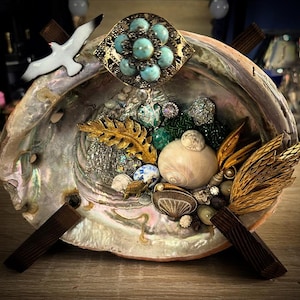 May include: Abalone shell art piece with a variety of decorative elements. Includes a vintage turquoise and gold brooch, seashells, gold-toned leaf and feather accents, and a white seagull figurine. The shell is mounted on a wooden base.