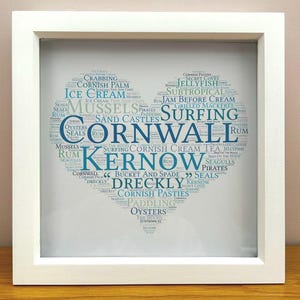 Può includere: Stampa con cornice bianca con una nuvola di parole a forma di cuore. Le parole, in blu e bianco, includono "Cornwall", "Kernow", "Surfing", "Ice Cream" e "Cornish Pasties". La stampa è su una superficie di legno.
