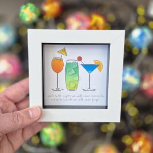 May include: A white framed art print with three cocktail illustrations. The drinks are orange, green, and blue, each with garnishes. The text reads, "Here's to the nights we will never remember and to the friends we will never forget."