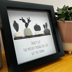 Op de afbeelding: Zwart omlijste kunstprint met vier gestileerde cactusontwerpen gemaakt van kiezelstenen. De print bevat de tekst "Don't let the prickly things in life get you down." Een decoratief item voor in huis.