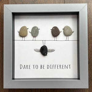 May include: A framed artwork featuring pebble art birds. Four birds perch on a wire, while a fifth, black bird hangs upside down below. The text "DARE TO BE DIFFERENT" is printed below the birds. The frame is gray.
