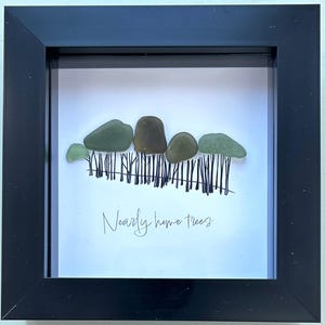 May include: A framed artwork featuring a minimalist landscape. The artwork depicts a row of trees made from smooth, rounded stones in shades of green and brown, with thin black lines representing the trunks. The text "Nearly home trees" is written below.