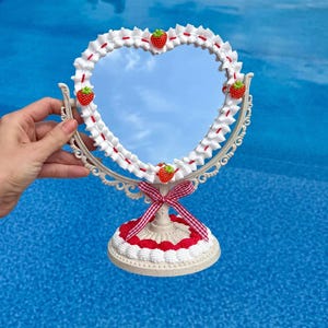 May include: A heart-shaped tabletop mirror with a cream-colored ornate frame. The mirror border is decorated with white whipped cream-like piping, small red strawberry charms, and a red and white checkered ribbon tied at the base of the stand.