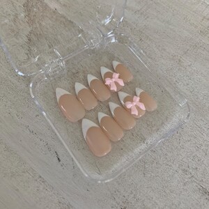 May include: A set of artificial nails in a clear plastic case. The nails have a nude base with white tips, resembling a French manicure. Two nails feature small pink bow accents. The nails are oval-shaped.