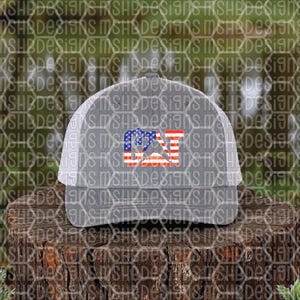 Puede incluir: Gorra de camionero gris con parte trasera de malla blanca y visera curva. La parte delantera presenta un dise&ntilde;o patri&oacute;tico con las letras "CAT" en rojo, blanco y azul, que recuerda a la bandera estadounidense. La gorra est&aacute; sobre una superficie de madera.