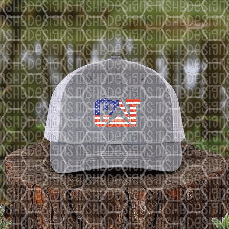 Puede incluir: Gorra de camionero gris con parte trasera de malla blanca y visera curva. La parte delantera presenta un dise&ntilde;o patri&oacute;tico con las letras "CAT" en rojo, blanco y azul, que recuerda a la bandera estadounidense. La gorra est&aacute; sobre una superficie de madera.