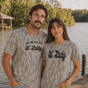 May include: Two people wearing matching gray t-shirts with the text "Let Me Ask My Ol' Lady" and "Ol' Lady" in black script. The shirts have a textured pattern. They are standing in front of a lake.