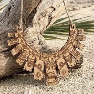 May include: A crescent-shaped wooden statement necklace featuring intricate laser-engraved tiki mask designs and geometric patterns. The piece is suspended from a thin, natural-colored twine cord, showcasing a light brown wood grain finish.