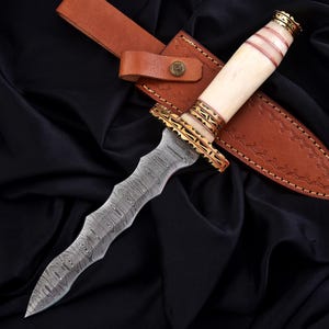 May include: A decorative dagger with a patterned steel blade and a bone handle accented with gold-colored metal. The dagger rests in a brown leather sheath with a matching belt loop.