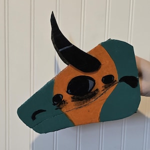 May include: A hand-painted cardboard cutout shaped like a dragon head in profile. The design features a teal and orange color scheme with a prominent black horn, a large black eye, and black painted details outlining the snout and facial features.