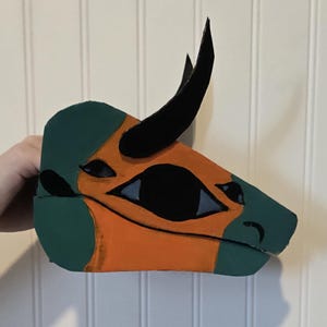 May include: A handcrafted dinosaur mask featuring a profile view of a head with a prominent black horn. The mask is painted in alternating sections of muted green and bright orange, with black accents around the eye and snout area.