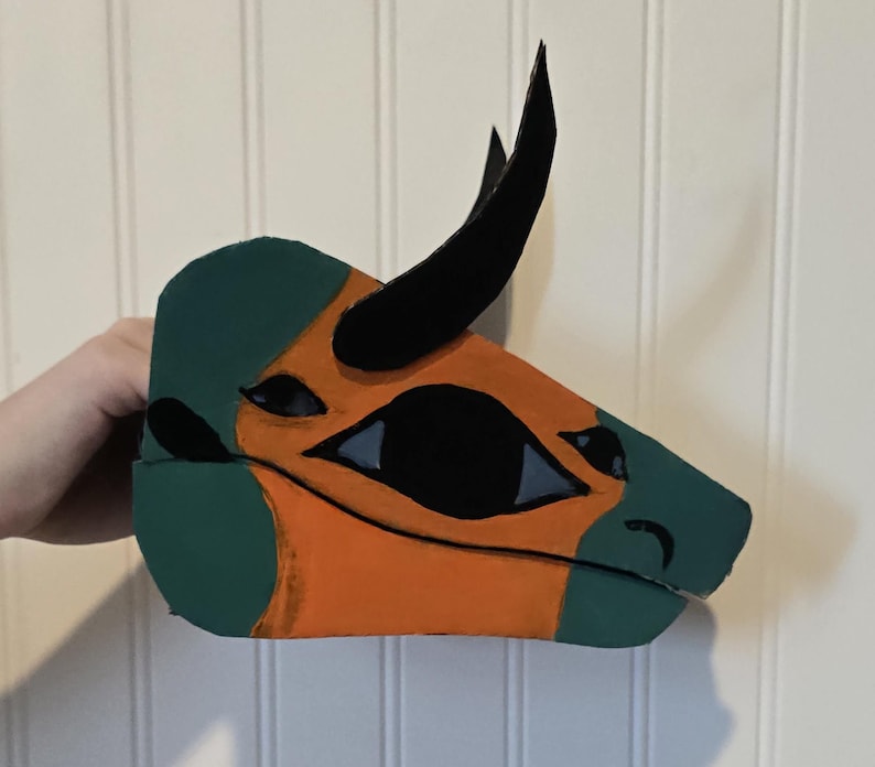 May include: A handcrafted dinosaur mask featuring a profile view of a head with a prominent black horn. The mask is painted in alternating sections of muted green and bright orange, with black accents around the eye and snout area.