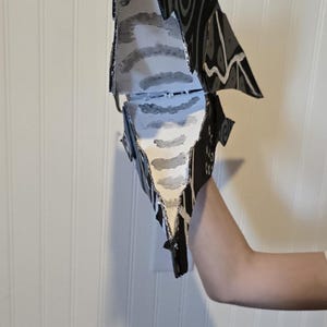 K&ouml;nnte beinhalten: Eine Handpuppe aus Pappe mit schwarzer Au&szlig;enseite, wei&szlig;en Wirbelmustern und einer wei&szlig;en Innenseite, die mit grauen horizontalen Streifen bemalt ist. Die Puppe hat eine spitze, kantige Form mit einem beweglichen Kiefermechanismus.