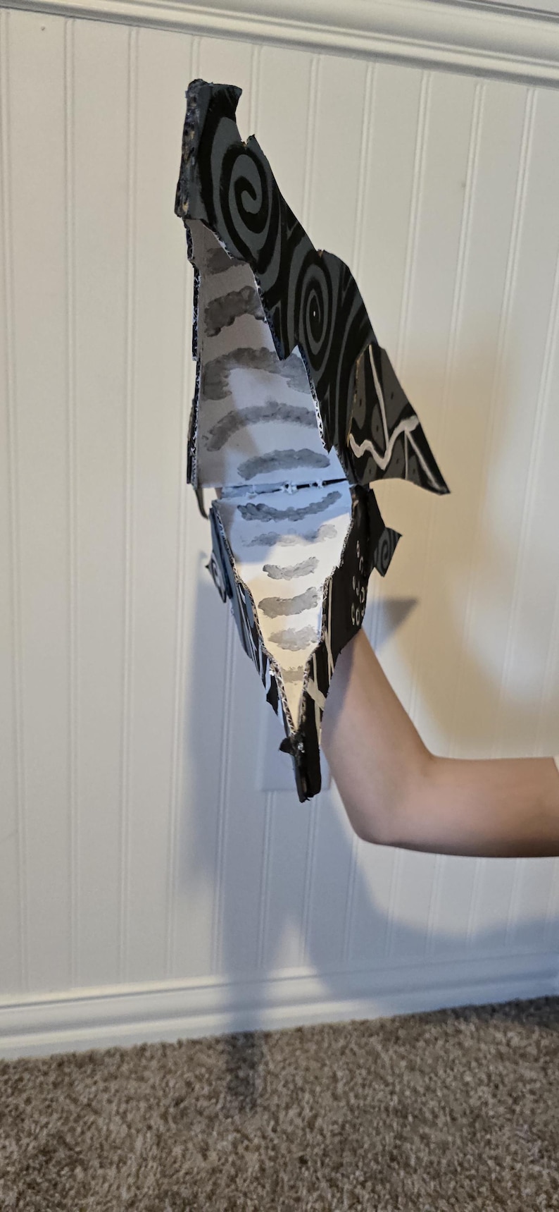 K&ouml;nnte beinhalten: Eine Handpuppe aus Pappe mit schwarzer Au&szlig;enseite, wei&szlig;en Wirbelmustern und einer wei&szlig;en Innenseite, die mit grauen horizontalen Streifen bemalt ist. Die Puppe hat eine spitze, kantige Form mit einem beweglichen Kiefermechanismus.
