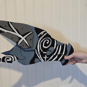 K&ouml;nnte beinhalten: Ein flaches, l&auml;ngliches und spitz zulaufendes Objekt mit einem bemalten Design in Schwarz-, Grau- und Wei&szlig;t&ouml;nen. Die Oberfl&auml;che zeigt abstrakte Muster, darunter wirbelnde Spiralen, scharfe winklige Linien und massive Farbfl&auml;chen.