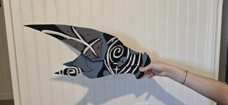 K&ouml;nnte beinhalten: Ein flaches, l&auml;ngliches und spitz zulaufendes Objekt mit einem bemalten Design in Schwarz-, Grau- und Wei&szlig;t&ouml;nen. Die Oberfl&auml;che zeigt abstrakte Muster, darunter wirbelnde Spiralen, scharfe winklige Linien und massive Farbfl&auml;chen.