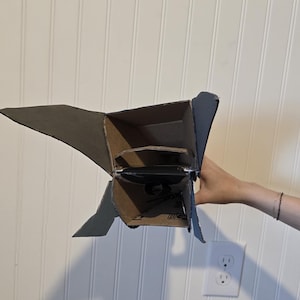 K&ouml;nnte beinhalten: Ein handgefertigtes Raketenmodell aus Pappe mit einem hohlen, rechteckigen K&ouml;rper und seitlich angebrachten dreieckigen Finnen. Die Struktur besteht aus brauner Wellpappe mit sichtbaren N&auml;hten und Kanten.
