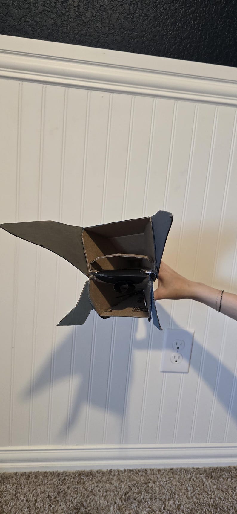 K&ouml;nnte beinhalten: Ein handgefertigtes Raketenmodell aus Pappe mit einem hohlen, rechteckigen K&ouml;rper und seitlich angebrachten dreieckigen Finnen. Die Struktur besteht aus brauner Wellpappe mit sichtbaren N&auml;hten und Kanten.