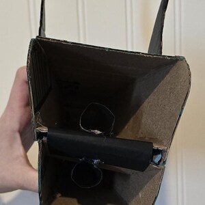 May include: A rectangular structure constructed from brown cardboard with black painted exterior edges. The interior features a horizontal black cardboard divider with two small circular openings, and two vertical black cardboard fins at the top.