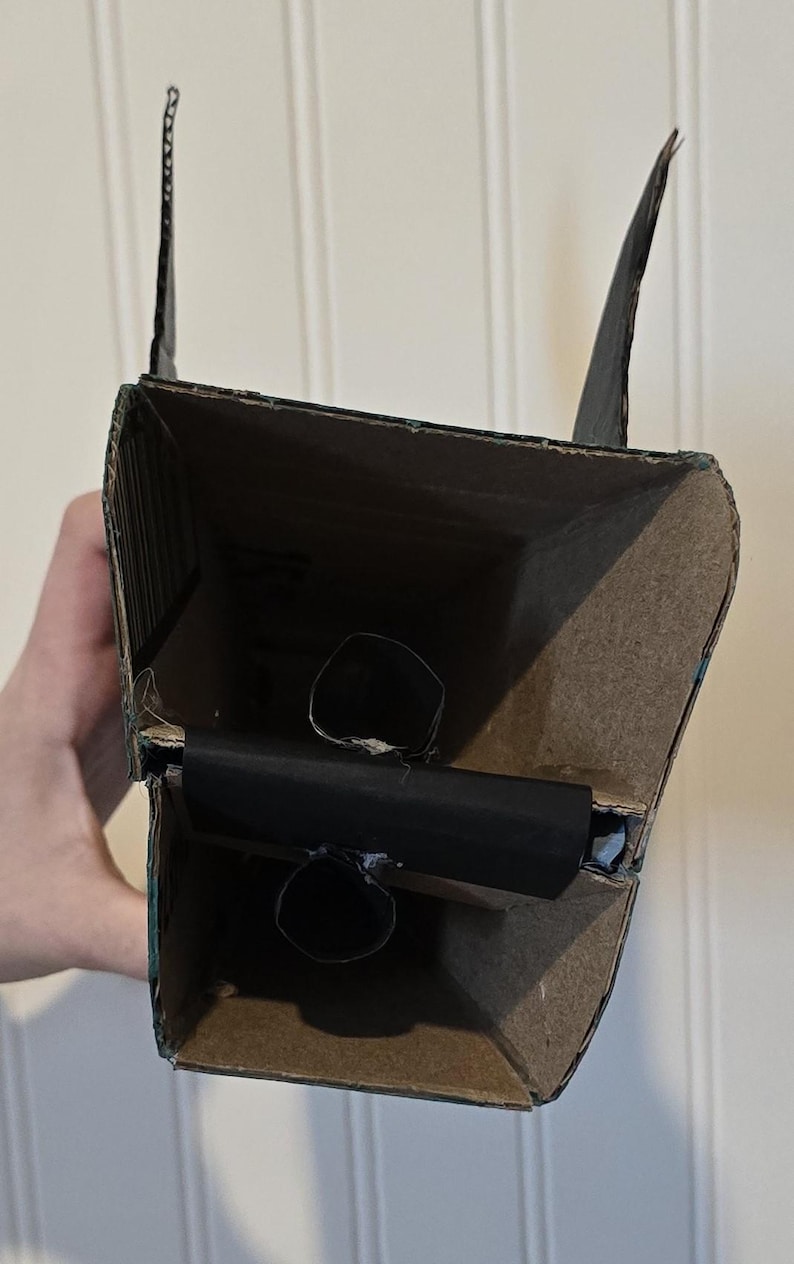 May include: A rectangular structure constructed from brown cardboard with black painted exterior edges. The interior features a horizontal black cardboard divider with two small circular openings, and two vertical black cardboard fins at the top.