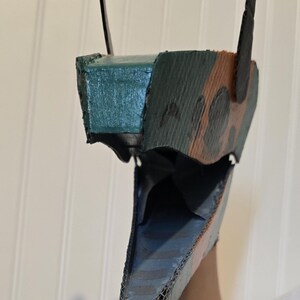May include: A handcrafted cardboard mask featuring a pointed snout and an open jaw. The exterior is painted with a camouflage pattern in shades of dark green, brown, and black, while the interior of the jaw is painted in a solid dark blue color.
