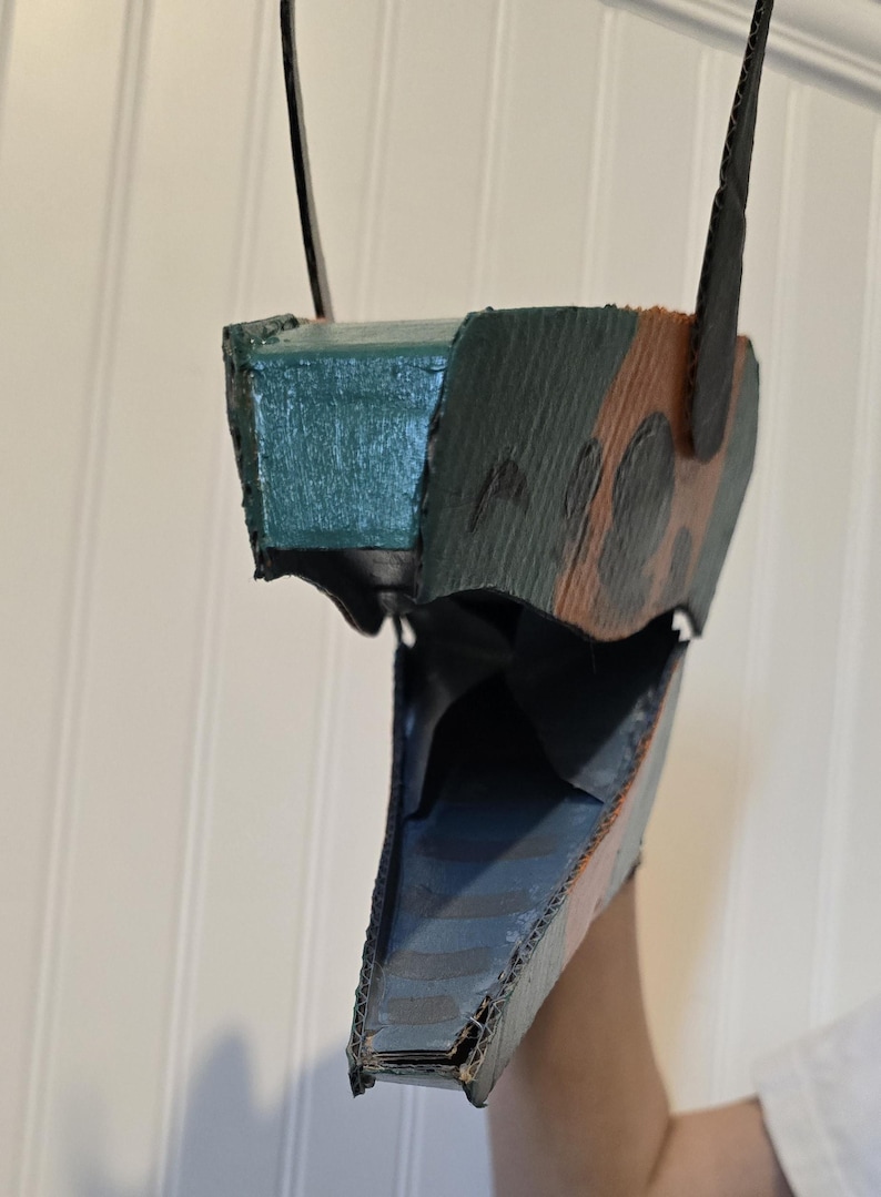 May include: A handcrafted cardboard mask featuring a pointed snout and an open jaw. The exterior is painted with a camouflage pattern in shades of dark green, brown, and black, while the interior of the jaw is painted in a solid dark blue color.