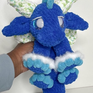 Puede incluir: Un dragón de peluche azul hecho a mano con ojos grandes y expresivos y pequeñas alas. El dragón presenta un material texturizado y suave y está acentuado con detalles blancos y azul claro. El peluche está diseñado con una apariencia caprichosa y amigable.