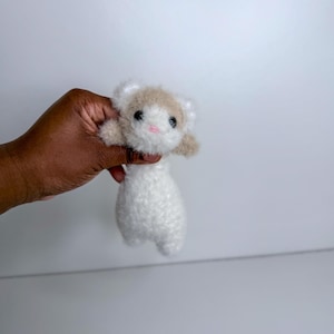 Op de afbeelding: Een handgemaakt, pluche fret speelgoed met een witte body en een kop met bruin en wit haar. De fret heeft zwarte ogen en een roze mond. Het speelgoed wordt in een hand gehouden tegen een witte achtergrond.