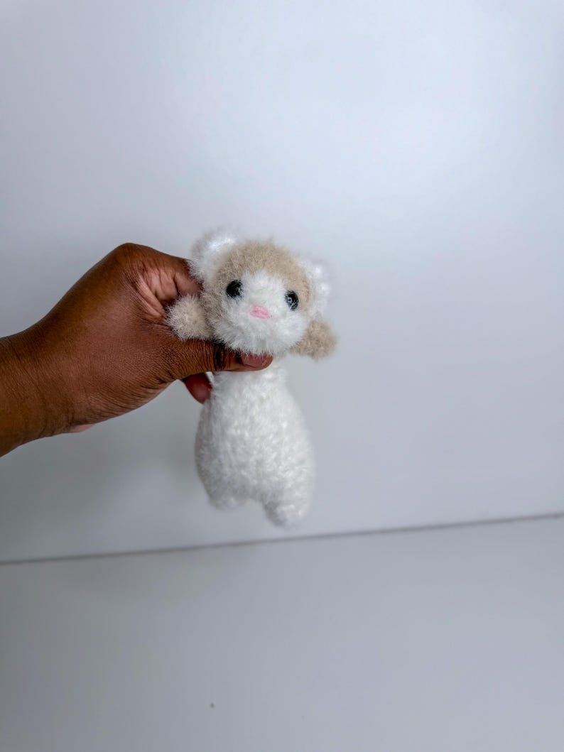 Op de afbeelding: Een handgemaakt, pluche fret speelgoed met een witte body en een kop met bruin en wit haar. De fret heeft zwarte ogen en een roze mond. Het speelgoed wordt in een hand gehouden tegen een witte achtergrond.