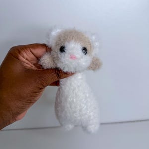 Op de afbeelding: Een handgemaakt pluche fret speelgoed met een witte body en een bruine kop. De fret heeft zwarte ogen en een roze neus. Het speelgoed is gemaakt van zacht, pluizig materiaal en wordt in een hand gehouden.