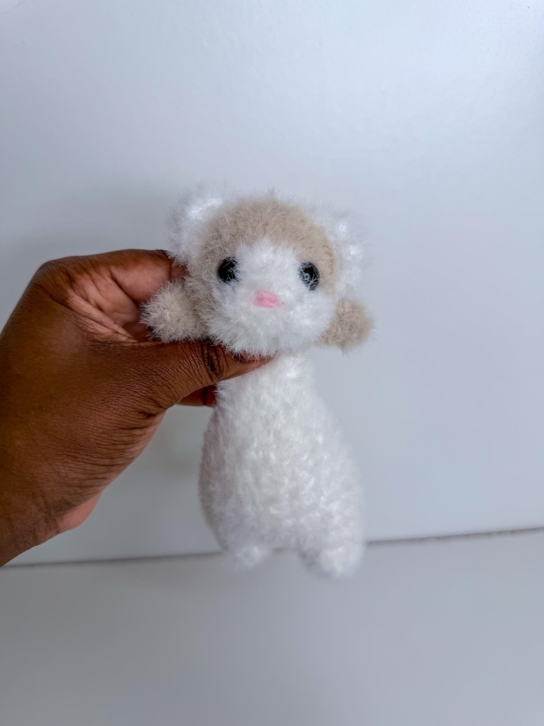 Op de afbeelding: Een handgemaakt pluche fret speelgoed met een witte body en een bruine kop. De fret heeft zwarte ogen en een roze neus. Het speelgoed is gemaakt van zacht, pluizig materiaal en wordt in een hand gehouden.