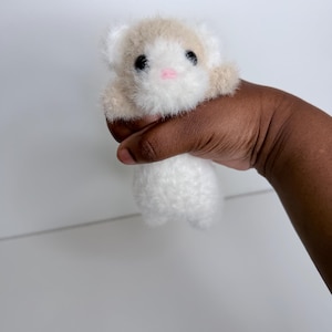 Op de afbeelding: Een klein, handgemaakt pluche speelgoed in de vorm van een hamster. Het speelgoed is wit met bruine accenten op het hoofd en de poten. Het heeft zwarte ogen en een roze neus. Het pluche wordt in een hand gehouden tegen een witte achtergrond.