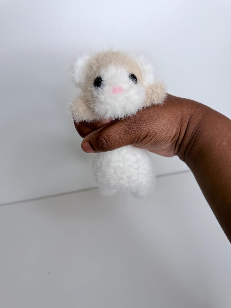 Op de afbeelding: Een klein, handgemaakt pluche speelgoed in de vorm van een hamster. Het speelgoed is wit met bruine accenten op het hoofd en de poten. Het heeft zwarte ogen en een roze neus. Het pluche wordt in een hand gehouden tegen een witte achtergrond.