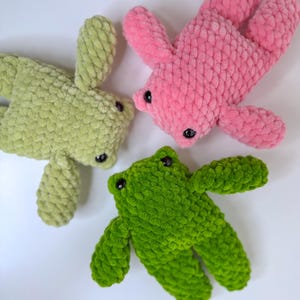 May include: Three plush frog toys made of soft, textured yarn. One frog is light green, one is a vibrant grass green, and one is bright pink. Each frog features small, round, black plastic eyes and a simple, rounded body shape with four limbs.