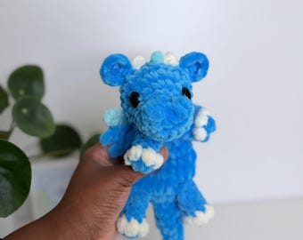 Puede incluir: Un dragón de peluche hecho de hilo azul brillante con un acabado suave y texturizado. La criatura presenta detalles blancos en las garras, las espinas de la espalda y la punta de la cola, junto con pequeñas alas azul claro y dos ojos negros.