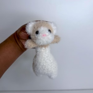 Op de afbeelding: Een handgemaakt knuffel ferretten speelgoed. De fret heeft een witte body, een bruine kop en kleine zwarte ogen. De neus van de fret is roze. Het speelgoed is gemaakt van zacht, pluizig materiaal.