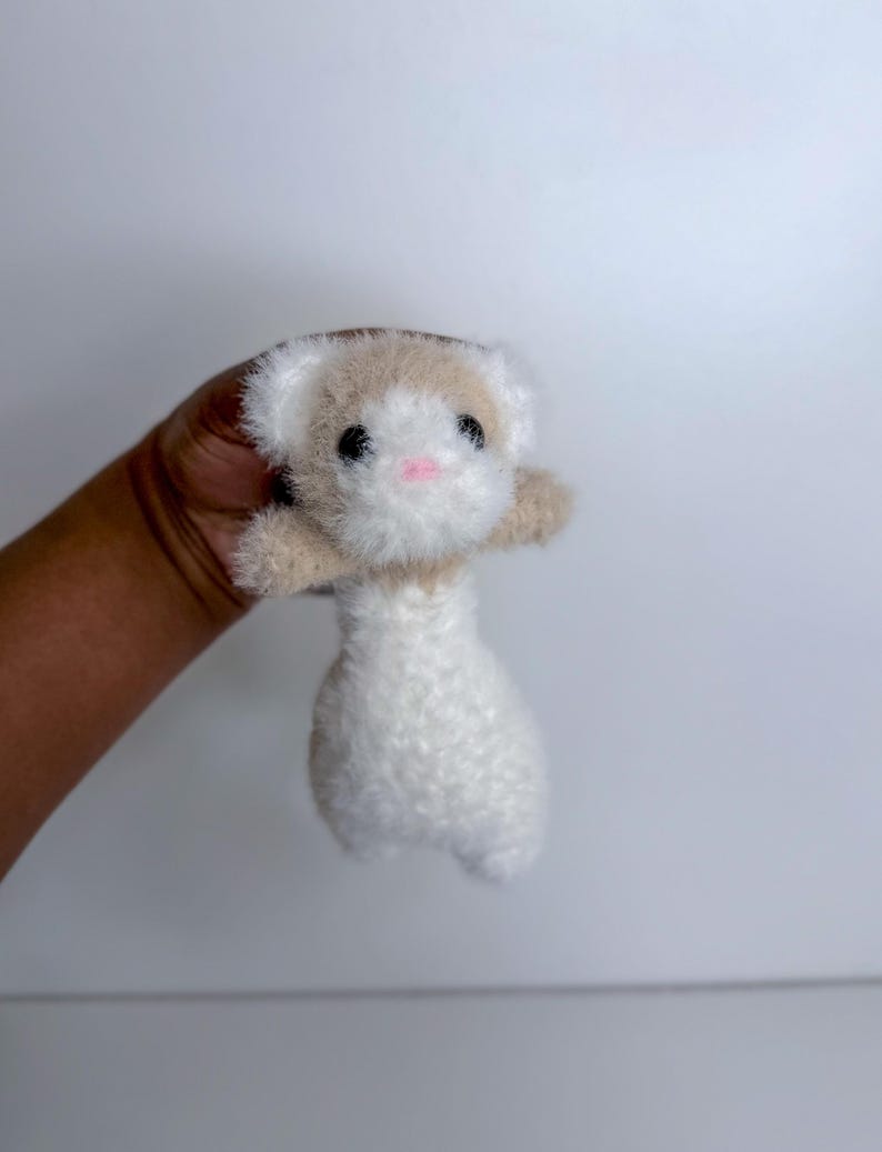 Op de afbeelding: Een handgemaakt knuffel ferretten speelgoed. De fret heeft een witte body, een bruine kop en kleine zwarte ogen. De neus van de fret is roze. Het speelgoed is gemaakt van zacht, pluizig materiaal.