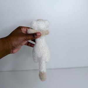 Op de afbeelding: Een klein, wit pluche speelgoed met een beige sjaal en een beige staart. Het speelgoed wordt tegen een witte achtergrond gehouden. Het pluche speelgoed is zacht en pluizig.