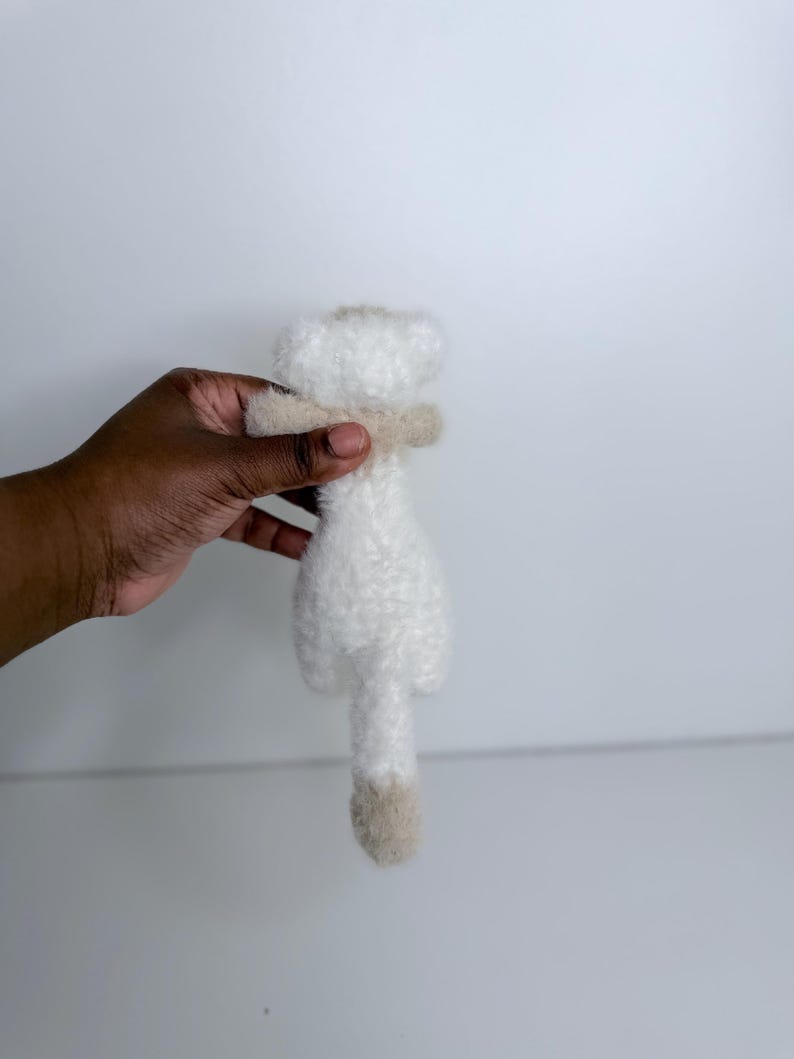 Op de afbeelding: Een klein, wit pluche speelgoed met een beige sjaal en een beige staart. Het speelgoed wordt tegen een witte achtergrond gehouden. Het pluche speelgoed is zacht en pluizig.
