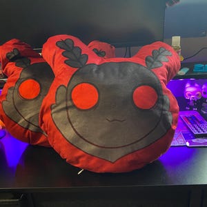 May include: Two plush pillows with a red and gray design. The pillows have a round face with large red eyes, a small mouth, and leaf-like accents. The pillows are on a black surface.