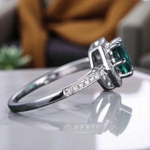 May include: A silver ring featuring a large, emerald-cut green gemstone. The ring has a halo of smaller, clear stones around the main gem and along the band. The ring is set on a reflective surface, showcasing the intricate design and sparkle.