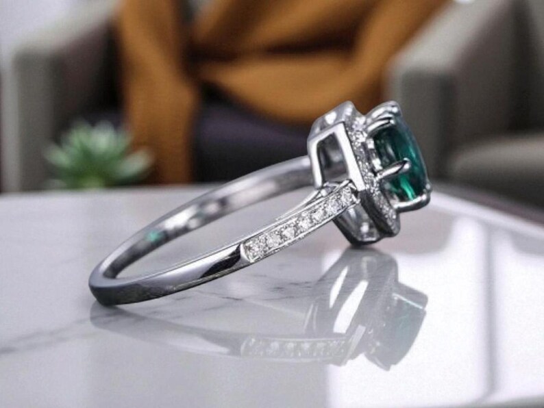 May include: A silver ring featuring a large, emerald-cut green gemstone. The ring has a halo of smaller, clear stones around the main gem and along the band. The ring is set on a reflective surface, showcasing the intricate design and sparkle.